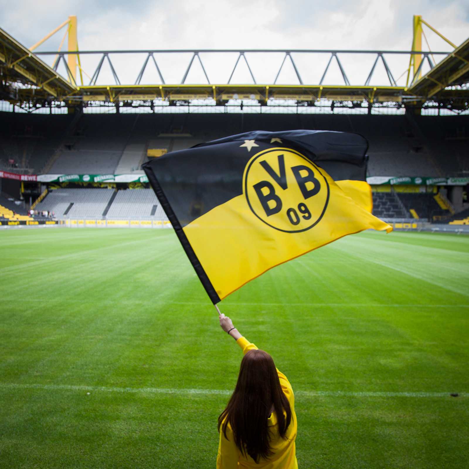 Person schwenkt die schwarzgelbe BVB-Stockfahne (90x60 cm) mit großem BVB-Emblem auf dem Rasen im SIGNAL IDUNA PARK, Rückansicht.