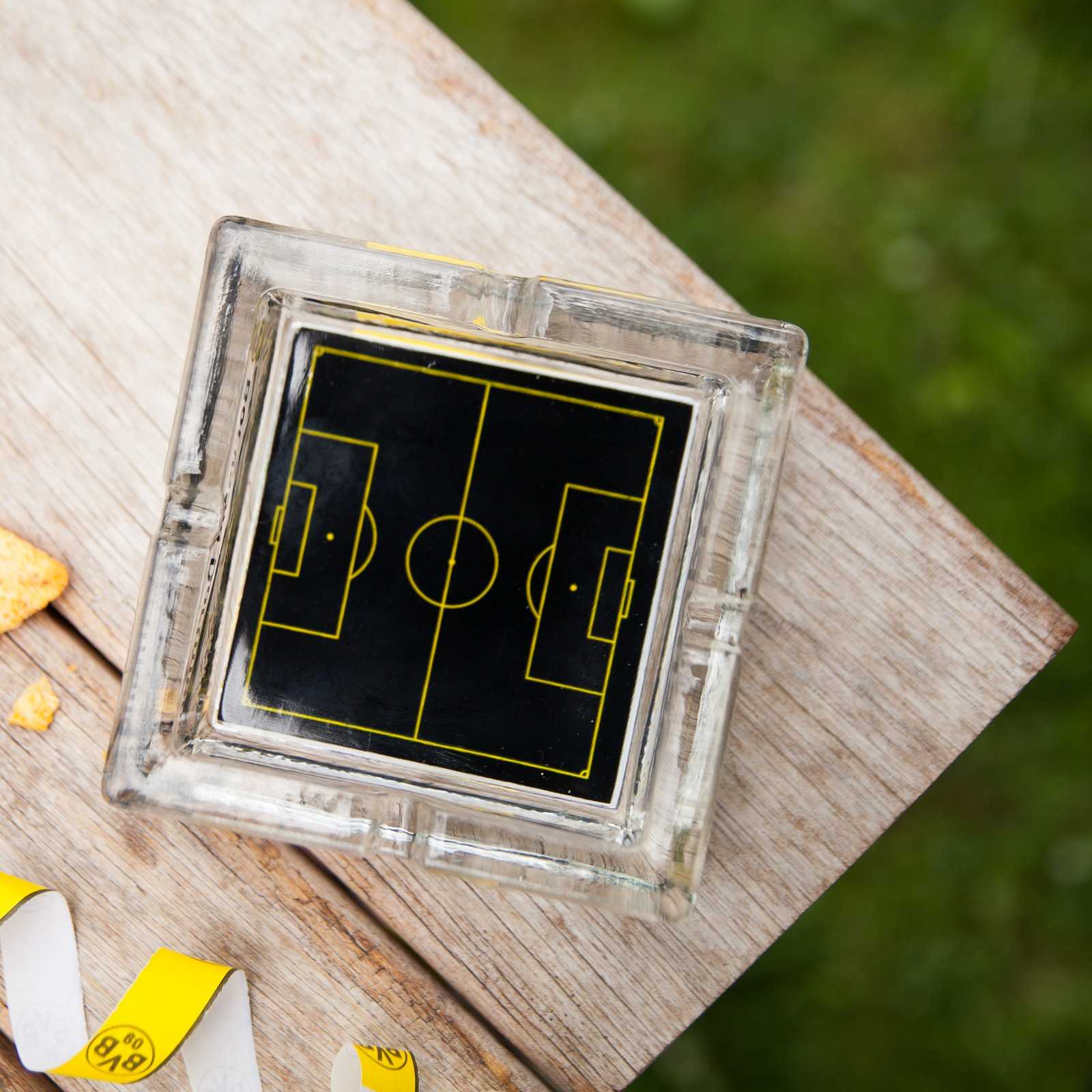 Detailansicht des BVB-Aschenbechers aus Glas mit schwarzgelbem Spielfeld-Design und BVB-Emblem, auf einem Holztisch platziert.