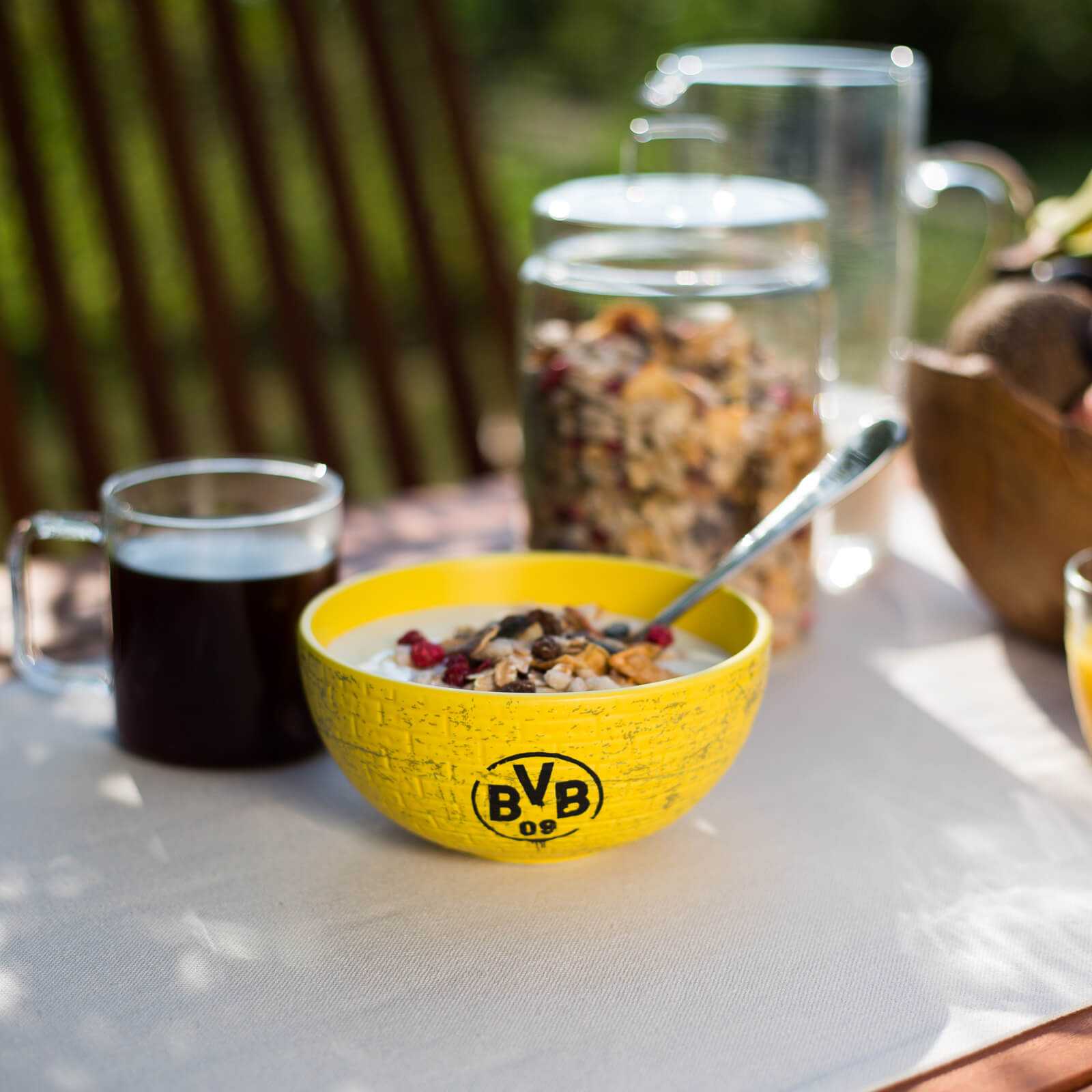 Nahaufnahme der BVB-Müslischale Gelbe Wand in leuchtendem Gelb mit BVB-Emblem, auf einem gedeckten Tisch und gefüllt mit Müsli.