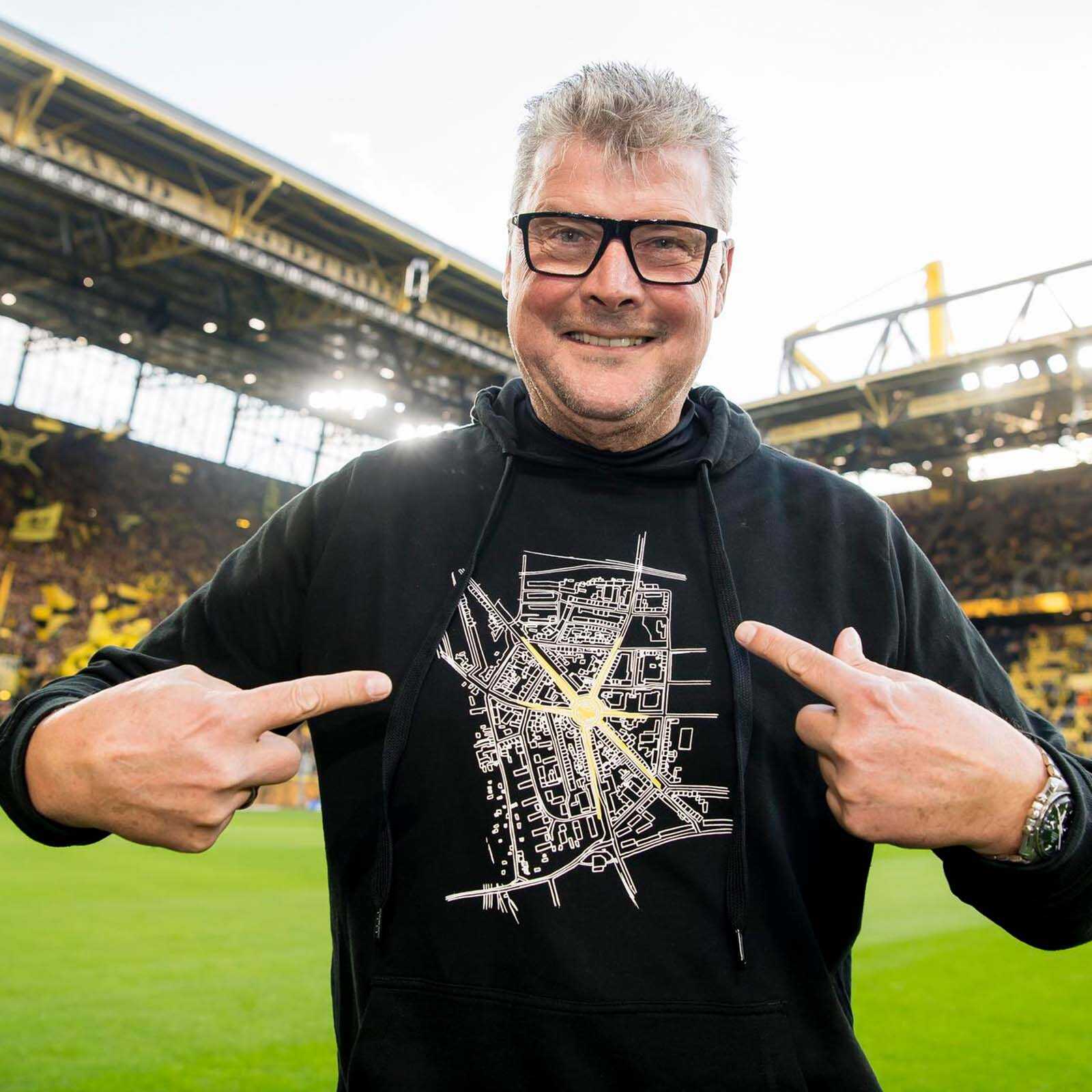 Ein Mann trägt den BVB Hoodie Leuchte auf, Unisex, in der Vorderansicht mit schwarzgelbem Stadtplan-Design und zeigt auf das Motiv. Im Hintergrund SIGNAL IDUNA PARK.