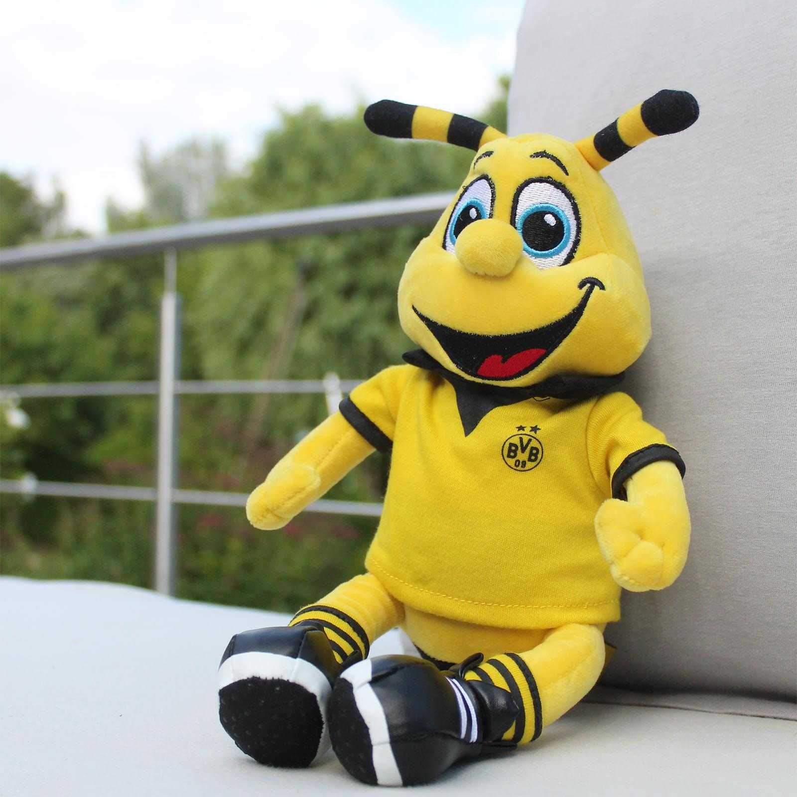 Vorderansicht der BVB-EMMA Plüschfigur mittel (30 cm) in schwarzgelbem Outfit mit BVB-Emblem auf dem T-Shirt, sitzend auf einem Sofa.