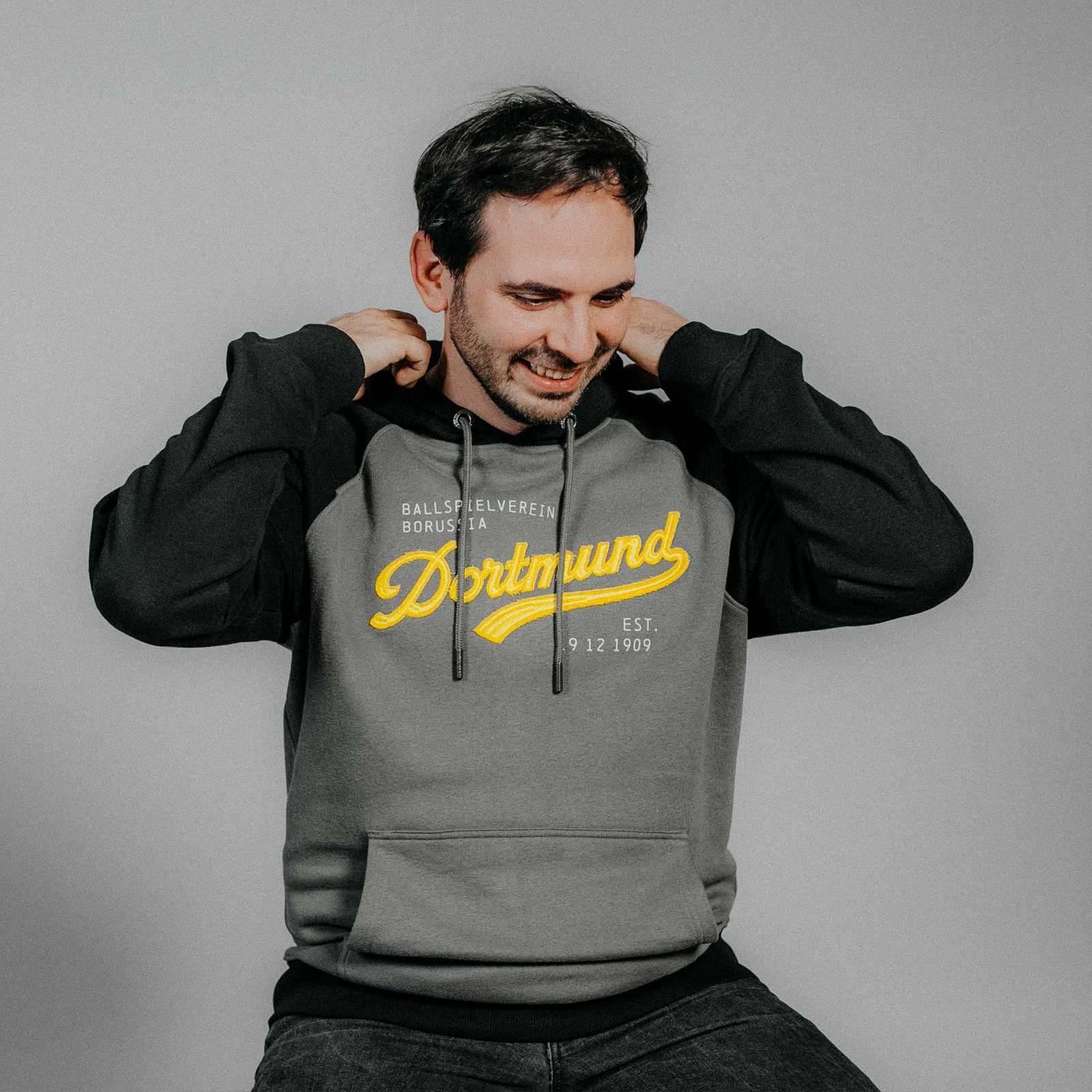 Ein Mann trägt den BVB Explorer Hoodie in der Vorderansicht; grau-schwarz mit gelbem "Dortmund"-Schriftzug und Kängurutasche, kein BVB-Emblem sichtbar.