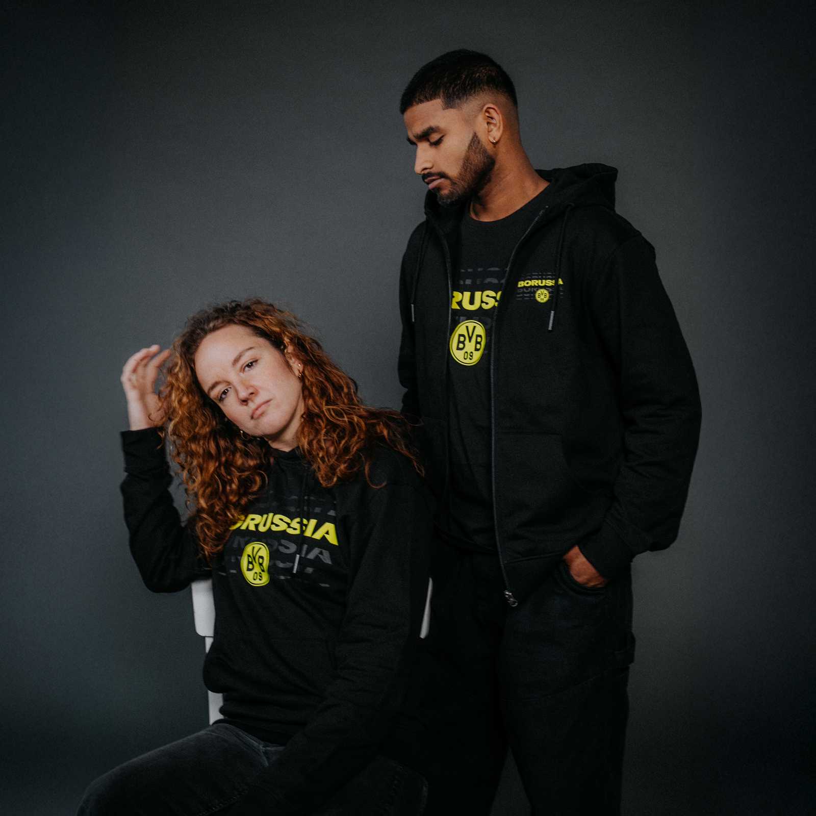 Zwei Personen tragen die BVB Sweatjacke black & neon in der Vorderansicht; beide Jacken sind schwarz mit neonfarbenem BVB-Emblem und Schriftzug.