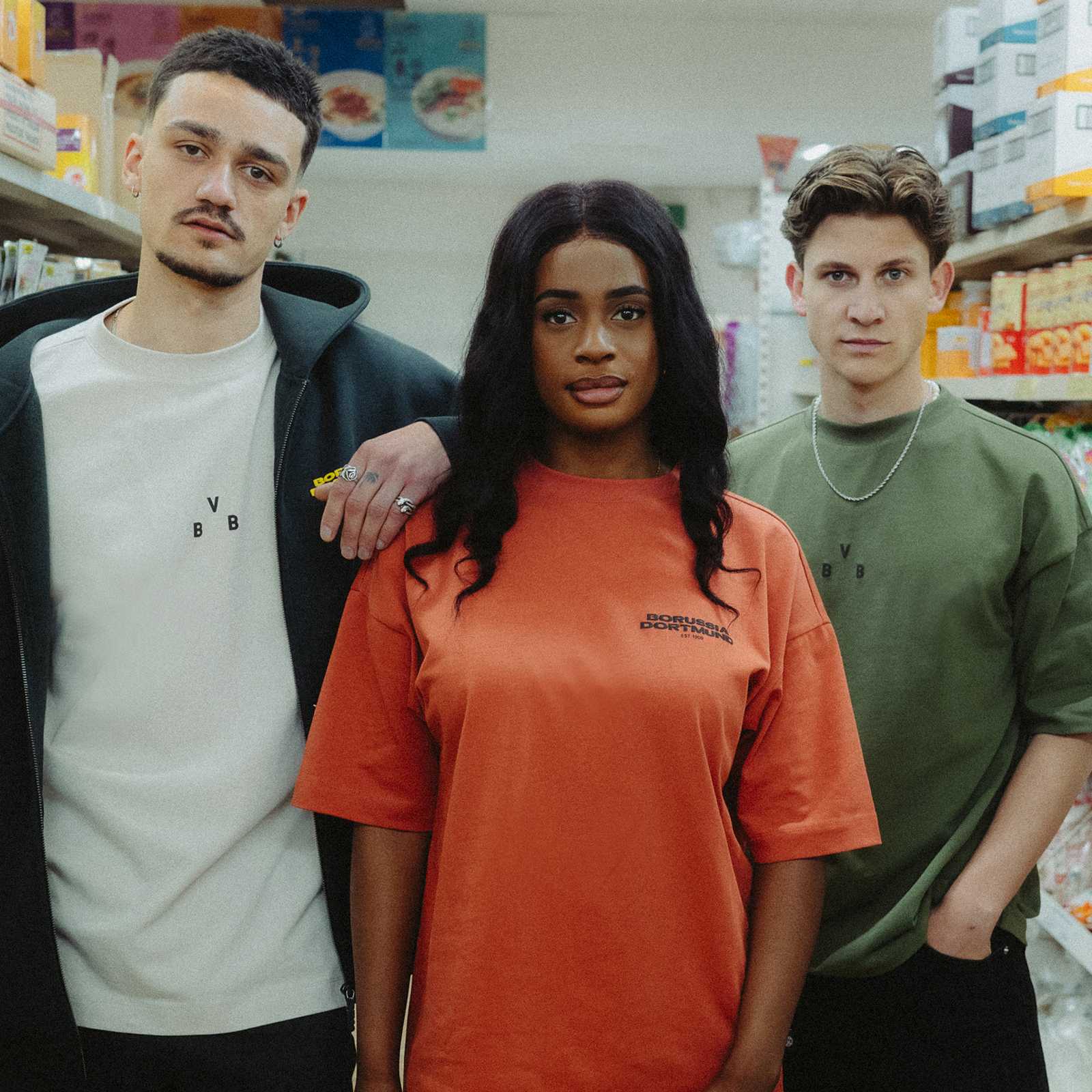 Drei Personen präsentieren in einer Vorderansicht das BVB Urban Tee orange 2025; das orange Shirt zeigt vorn den „Borussia Dortmund“-Schriftzug.