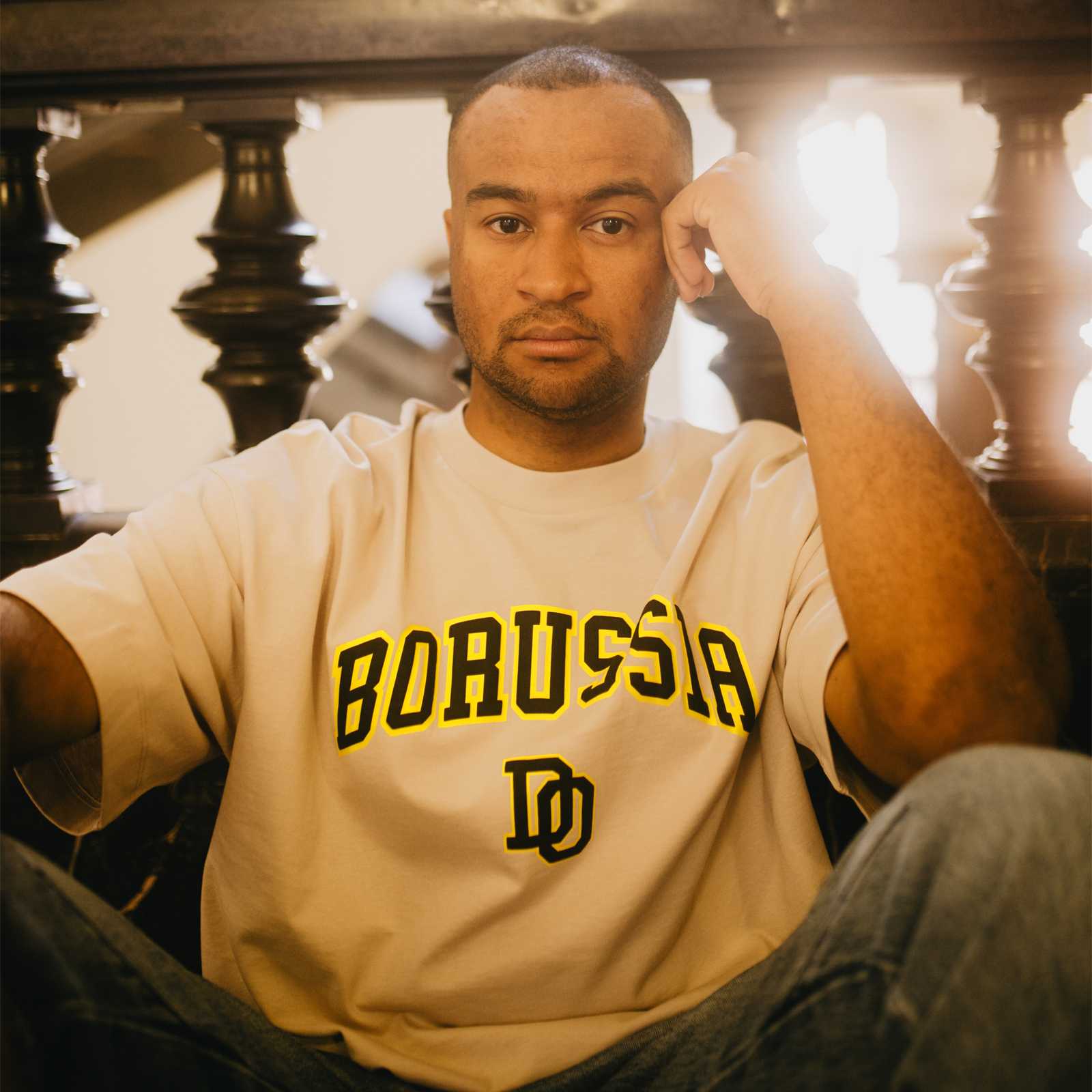 Frontalansicht eines Mannes im beige BVB College T-Shirt mit schwarzgelbem "BORUSSIA" Schriftzug und BVB-Initialen, der das Shirt trägt.