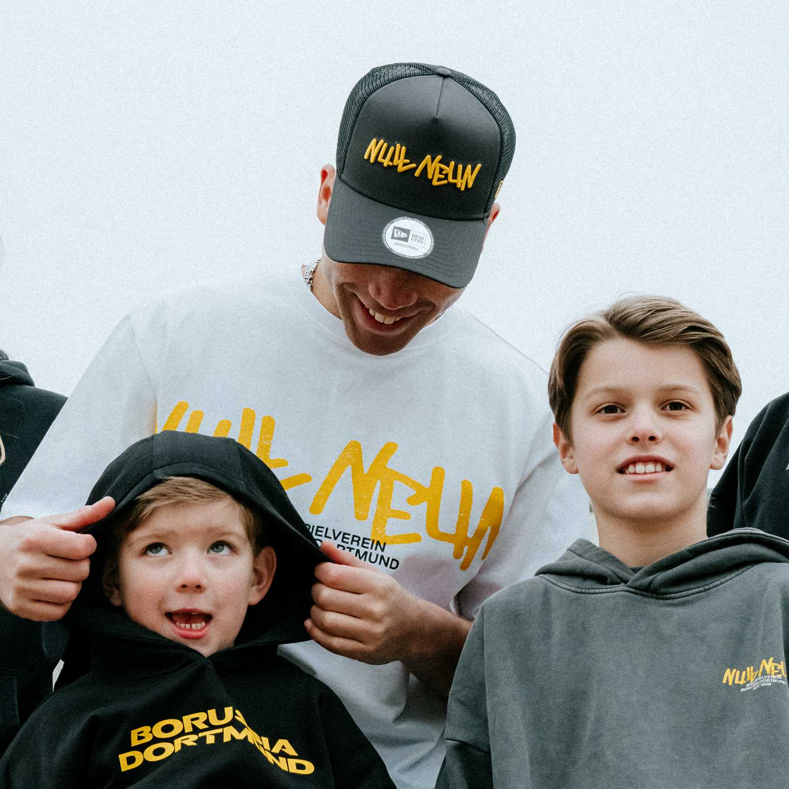 Ein Mann im weißen Shirt trägt die graue BVB-Kappe und steht hinter zwei jungen die BVB-Pullis tragen.
