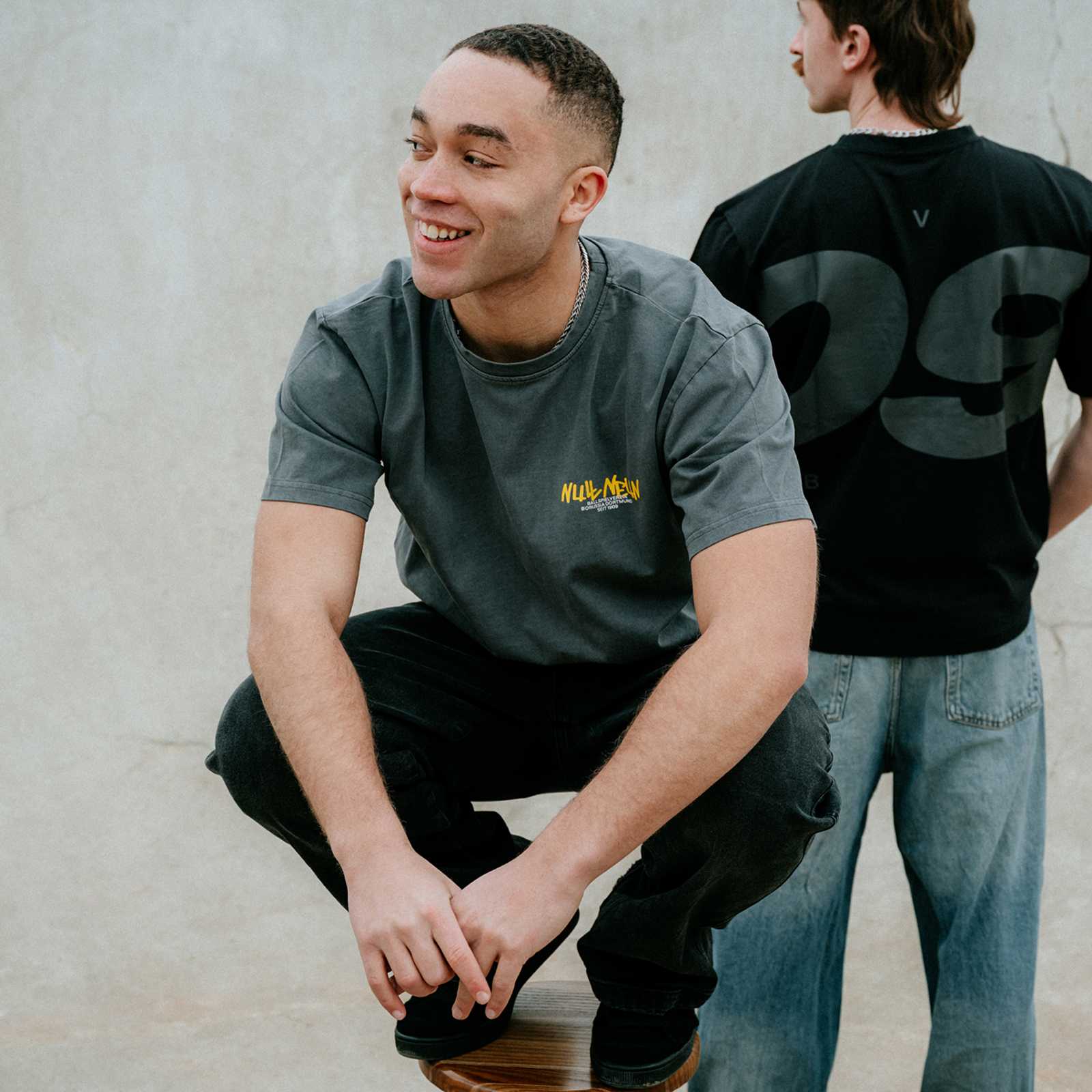 Ein männliches dunkelhaariges Model sitzt auf einem Hocker und trägt das T-Shirt.