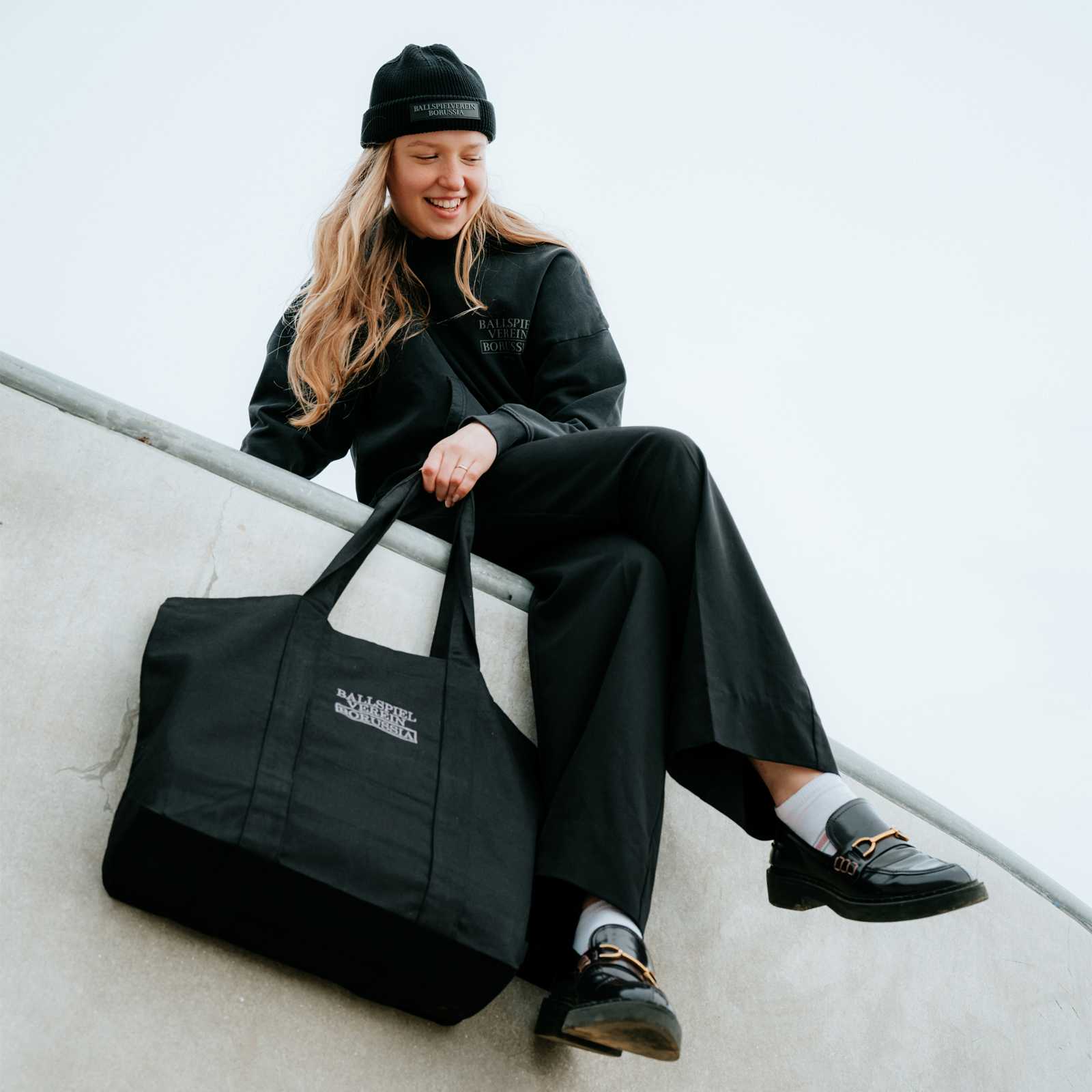 Ein weibliches blondes Model sitzt auf dem Rand einer Skate-Bahn und hält die Tasche in der Hand