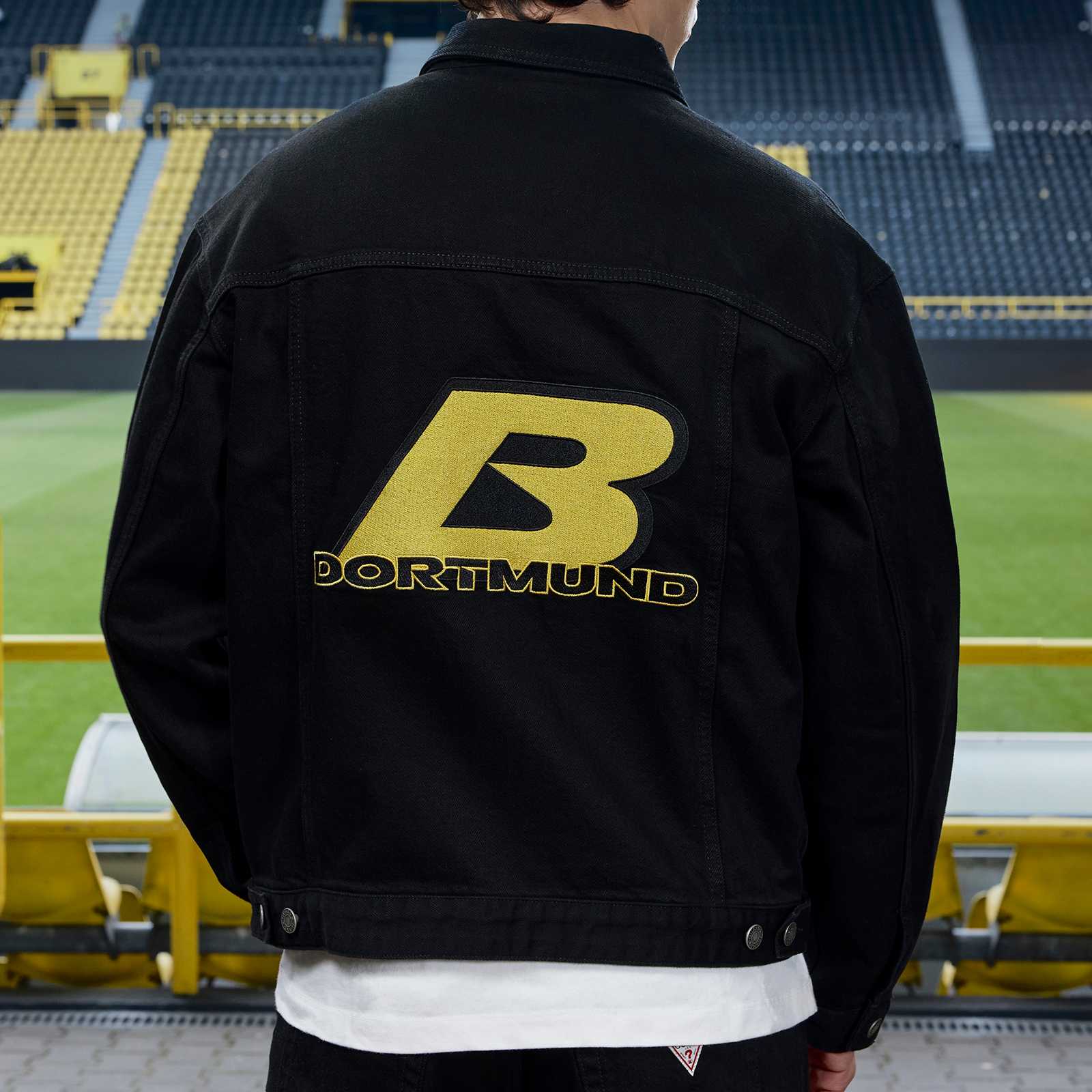 Junges männliches Model steht im Stadion mit dem Rücken zur Kamera und trägt die schwarzen BVB-Jeansjacke mit großem B und Dortmund Print auf dem Rücken.