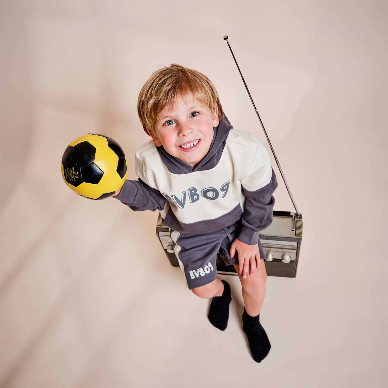 Kleiner Junge sitzt auf einem alten Radio und schaut nach oben in die Kamera. Er trägt den bei-grauen BVB-Hoodie mit passender Shorts und hält einen kleinen, schwarzgelben Ball in der Hand.