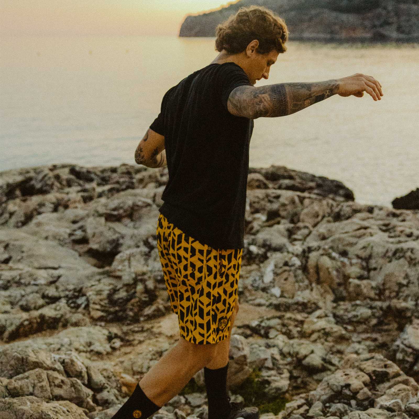 Ein männliches Model mit schwarzem T-Shirt trägt die Shorts und läuft über Felsen am Strand