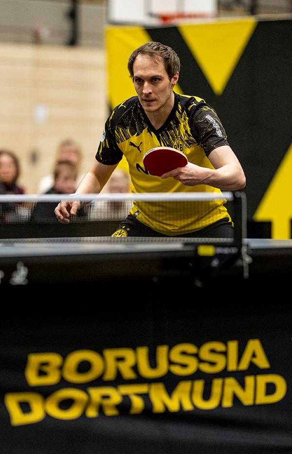 BVB Tischtennisspieler im Heimtrikot steht an der Tischtennisplatte und hält den Schläger hoch.